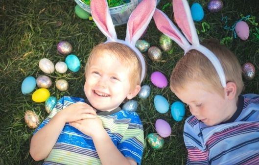 Two laughing young children laying on the grass wearing Easter bunny ears headbands, and with painted Easter eggs around them on the grass.