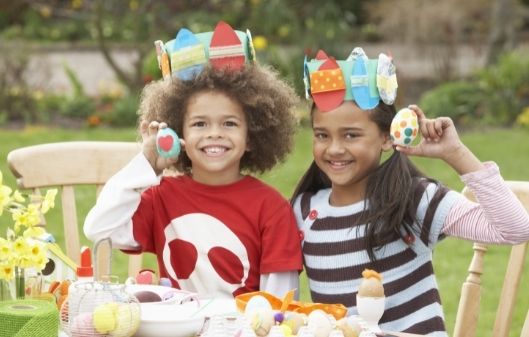 Two young children pictured at an Easter party, wearing party hats and showing off their painted Easter eggs.