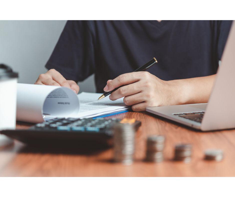 person sat at desk with coins stacked, laptop open and pen in hand