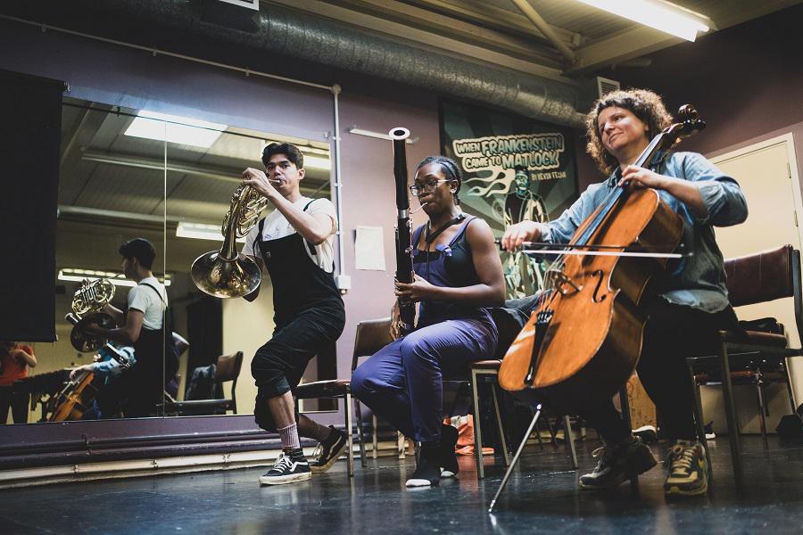 Alt text: A person playing cello seated. A person playing bassoon seated and a person playing French horn standing