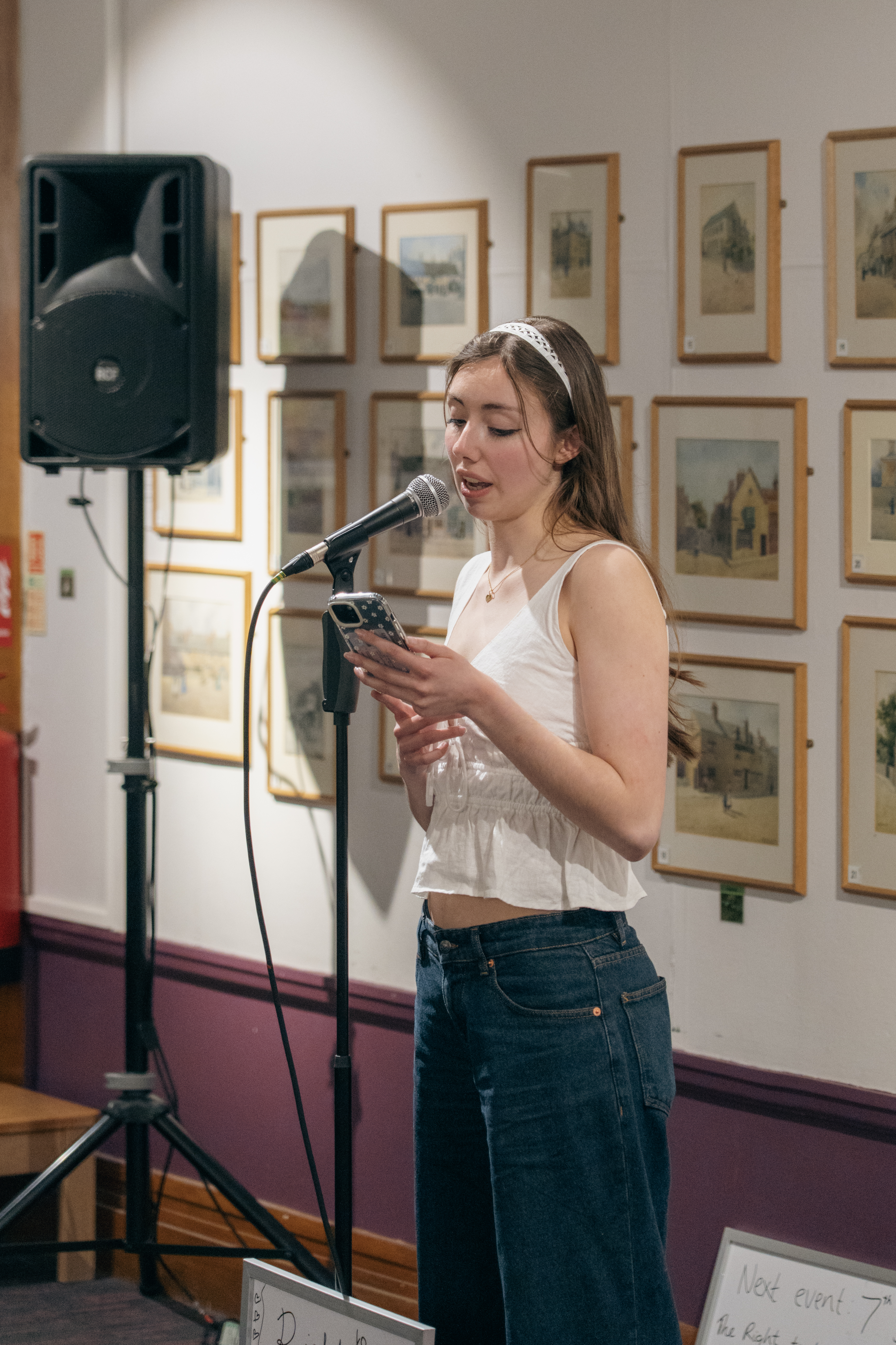 An Image of a white woman with brown hair speaking into a microphone. She is wearing a white top and dark blue jeans. She is reading from her mobile phone. Behind her are a selection of painting in a gallery space