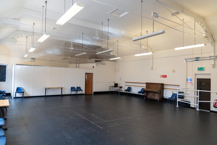 a large room with white walls, high apex ceiling  and dark floor. In the room is an upright piano and a slection of chairs