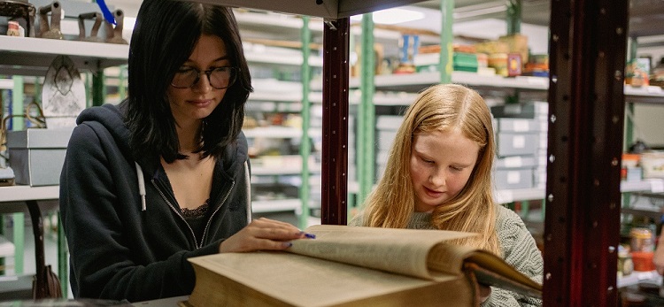 Two girls are leafing through a large book on a shelf in a museum store, while other historic objects can be seen on shelves in the background