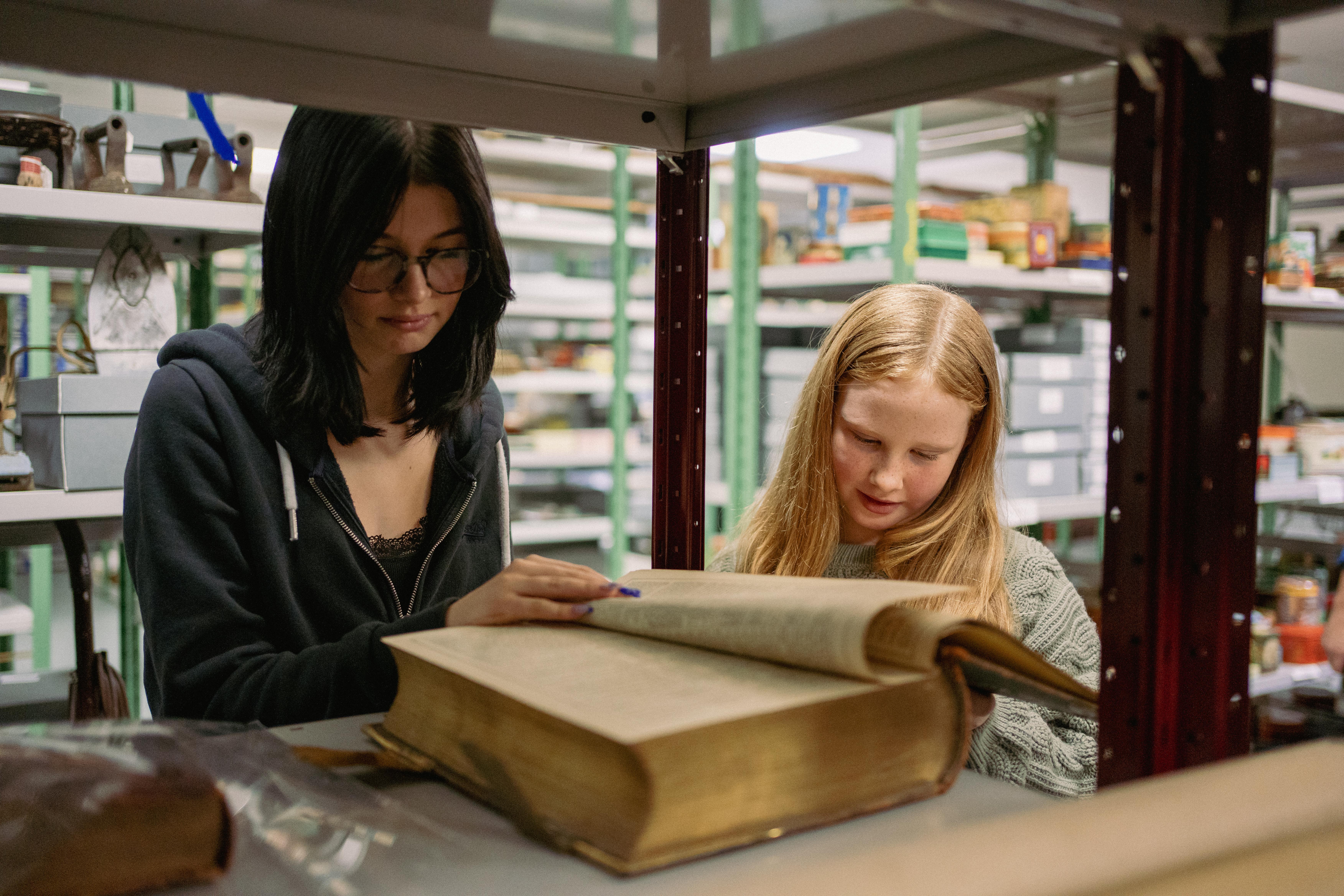 two young females looking at a large, old book in a Museum store, surrounded by shelves of objects