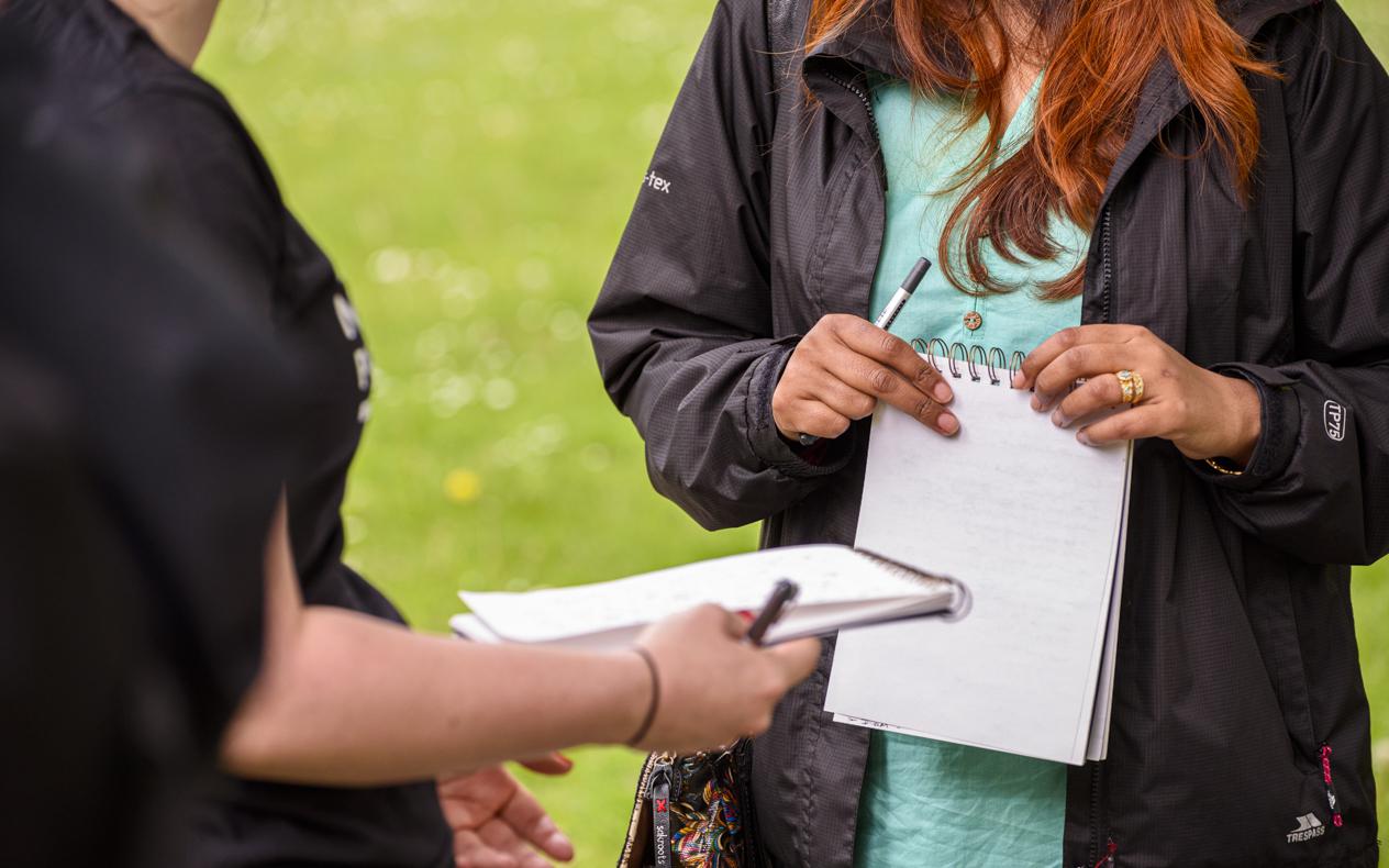 two people , not showing faces, outside with green field in background with pens and notebooks in hands