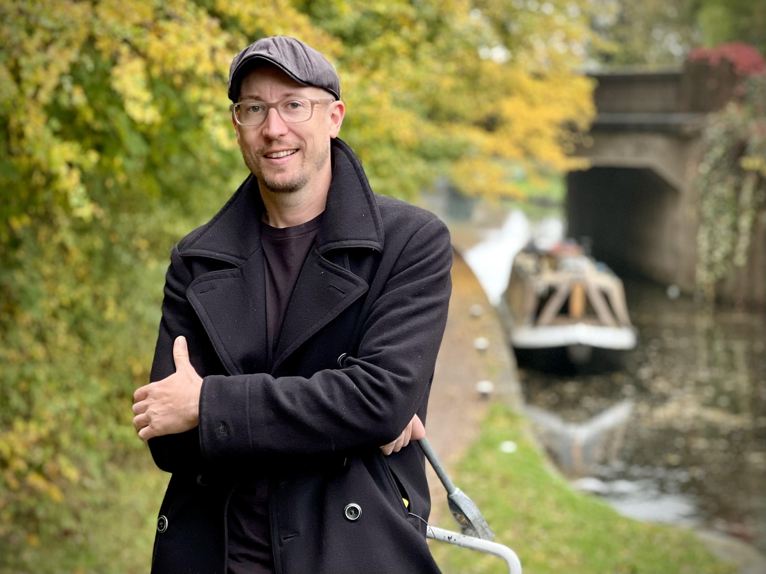 man in a blue coat, glasses and flat cap standing smiling with arms folded. In background autumn trees, canal and boat.